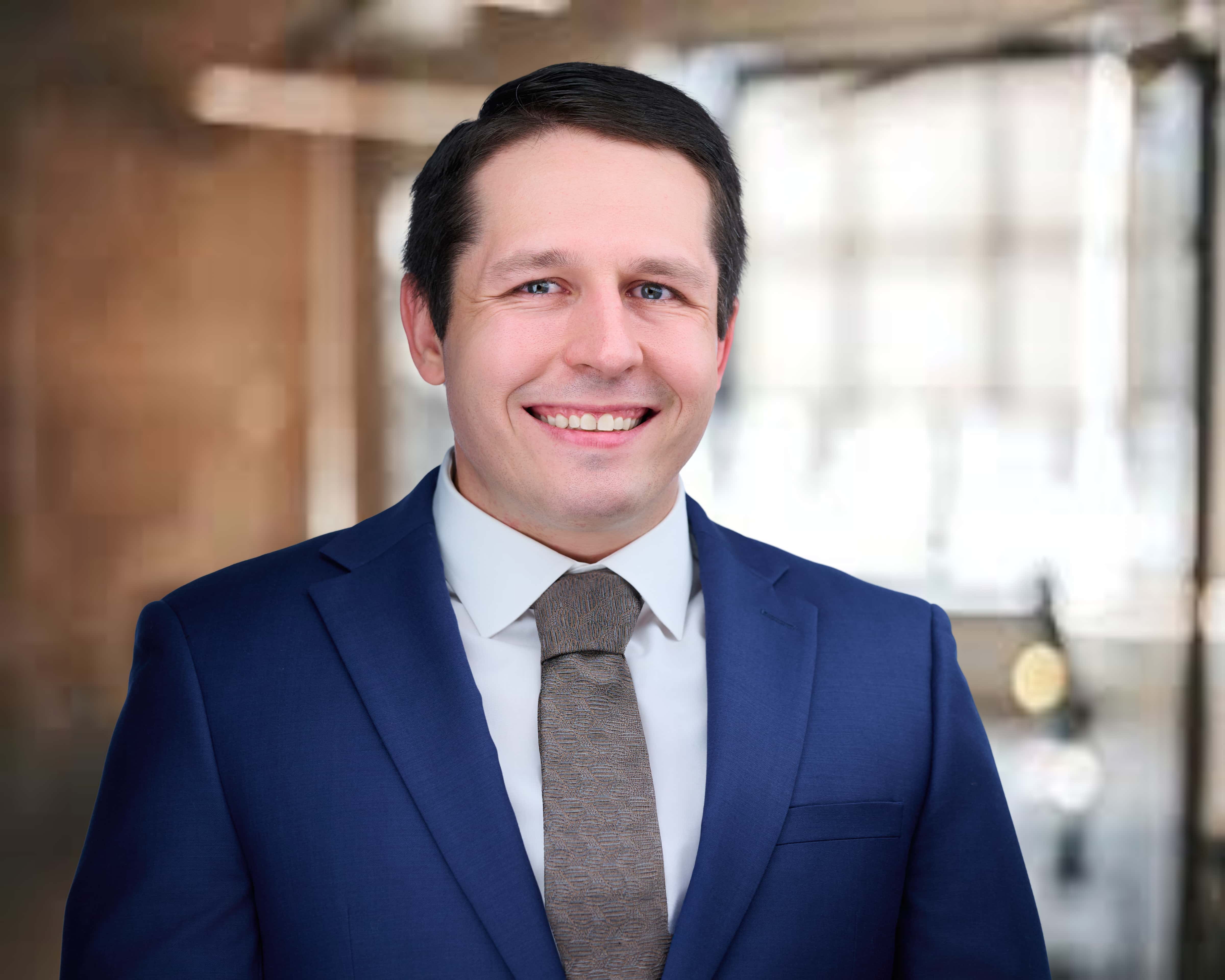 Smiling man with dark hair wearing a blue suit and brown tie in a blurred indoor office setting.