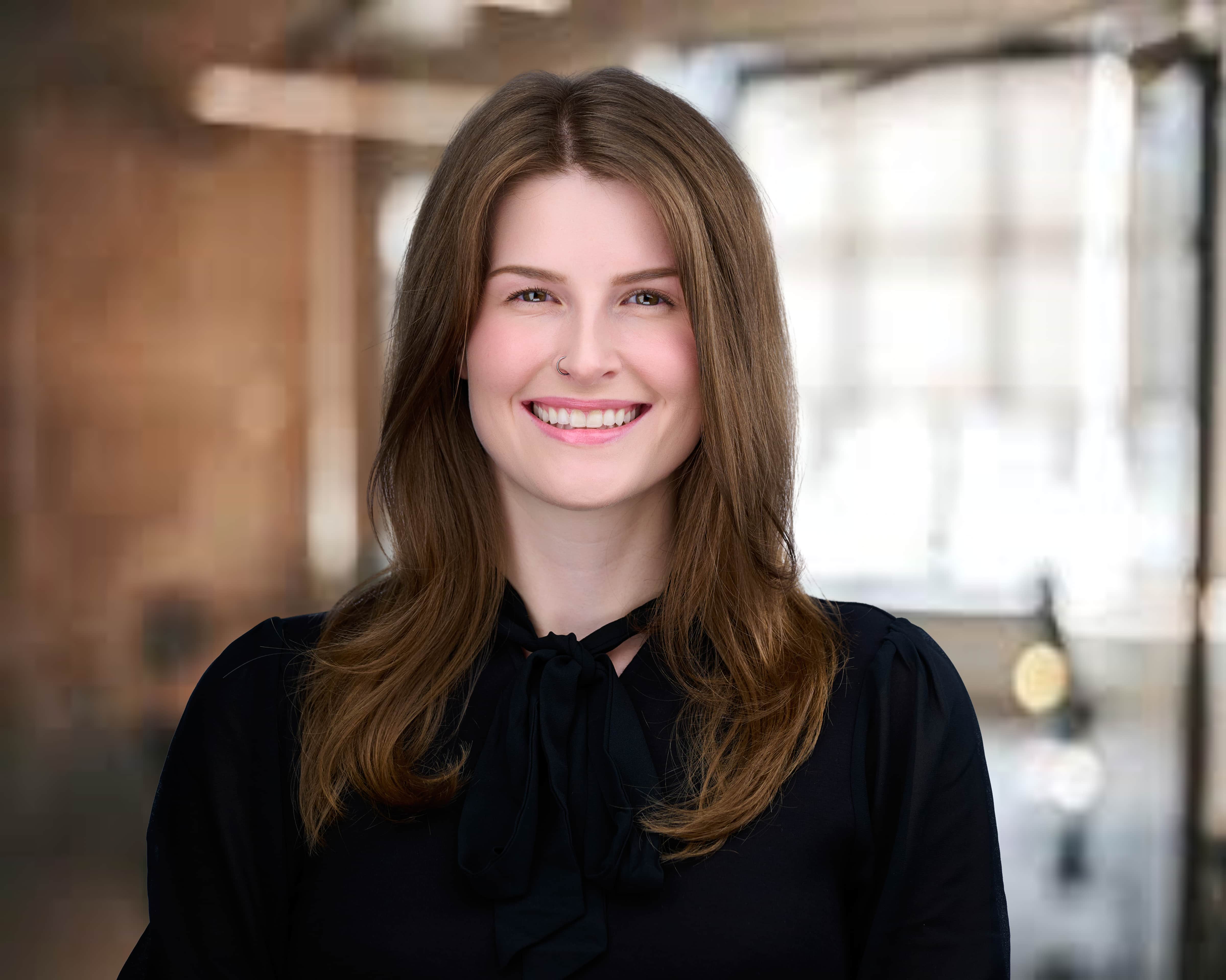 Smiling young woman with long brown hair wearing a black top with a bow in a softly blurred office background.